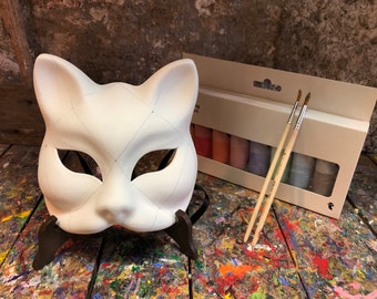May include: A white cat-shaped mask with black lines, resting on a brown stand. A set of paint tubes and two paintbrushes are next to the mask. The mask is ready to be painted.