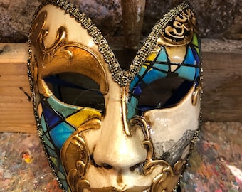 May include: A white and gold Venetian masquerade mask with blue and yellow stained glass accents. The mask has a smiling face and the word "Carnevale" written on the side.