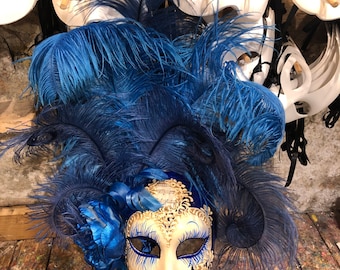 Può includere: Una maschera bianca da carnevale con dettagli blu e dorati e un pennacchio di piume blu. La maschera ha un design delicato ed è perfetta per un ballo in maschera o una festa in costume.