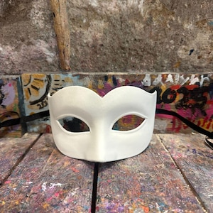 May include: A plain white masquerade mask with eye cutouts. The mask is made of a smooth material and is designed to cover the upper face. It rests on a wooden surface with colorful paint splatters.
