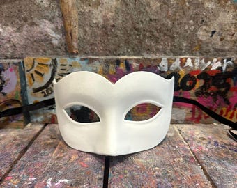 Può includere: Una maschera bianca semplice con fori per gli occhi. La maschera è realizzata in un materiale liscio ed è progettata per coprire la parte superiore del viso. Poggia su una superficie di legno con schizzi di vernice colorati.