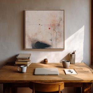 May include: An abstract painting in a light wood frame hangs above a wooden desk. The painting features a beige background with black and red accents. A laptop, mug, and books sit on the desk, along with a pencil holder and a small notepad.