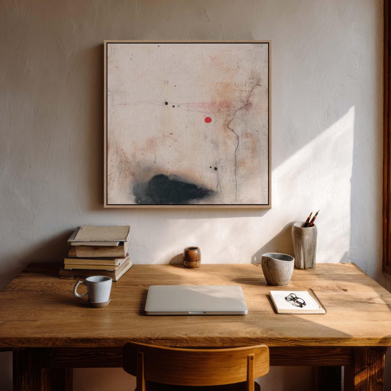 May include: An abstract painting in a light wood frame hangs above a wooden desk. The painting features a beige background with black and red accents. A laptop, mug, and books sit on the desk, along with a pencil holder and a small notepad.
