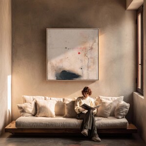 May include: A person reading on a beige sofa with several pillows. Above the sofa is a framed abstract painting with a red dot and black shapes. The room has a neutral color palette with natural light.