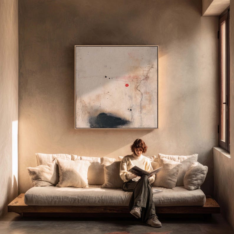 May include: A person reading on a beige sofa with several pillows. Above the sofa is a framed abstract painting with a red dot and black shapes. The room has a neutral color palette with natural light.