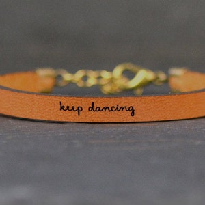 May include: A brown leather bracelet with a gold clasp and the words "keep dancing" engraved on the front.