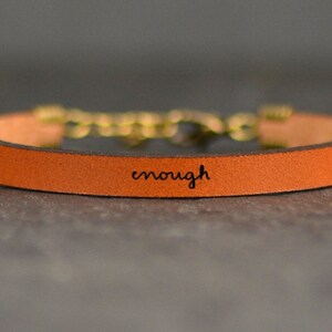 May include: A brown leather bracelet with a gold clasp and the word "enough" engraved on the band.