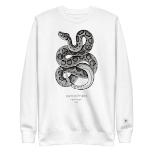Può includere: Felpa bianca con un'illustrazione in bianco e nero di un serpente. Il testo "Darwin Project", "Light Bringer" e "S24" è stampato sotto il serpente.
