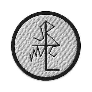 May include: Embroidered patch with a circular design. The patch has a light grey background and a black border. A black symbol is embroidered in the centre, resembling a stylized compass or emblem.