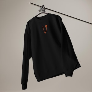 Pode incluir: Camisola preta de mangas compridas, pendurada num cabide de metal. A camisola apresenta um design de alfinete de segurança bordado a laranja na parte superior do peito. A peça de vestuário é feita de um material macio e confortável.