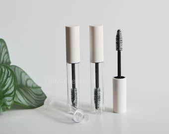 May include: Two clear plastic mascara tubes with white caps and a black mascara wand. The tubes are on a white surface with a green plant in the background.