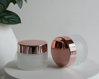 May include: Two frosted glass jars with rose gold lids and bands. The jars are cylindrical and sit on a white surface. The lids are shiny, reflecting light. The background is a soft white with a hint of a green plant.