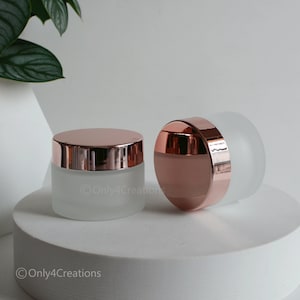 May include: Two frosted glass jars with rose gold lids and bands. The jars are cylindrical and sit on a white surface. The lids are shiny, reflecting light. The background is a soft white with a hint of a green plant.