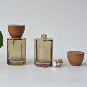 May include: Two hexagonal glass perfume bottles with a light brown tint. One bottle is capped with a rounded wooden lid, while the other stands open with its silver spray nozzle exposed and its matching wooden cap placed beside it on the surface.