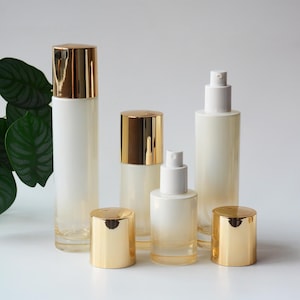 Puede incluir: Un conjunto de cinco envases cosméticos con cuerpos de vidrio blanco y tapas doradas reflectantes. El set incluye dos botellas altas con tapas doradas, una botella mediana con tapa dorada y dos botellas más pequeñas con dispensadores blancos.