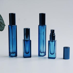 May include: Four rectangular glass spray bottles in a deep blue hue. Two tall bottles and one shorter bottle are topped with dark caps. One shorter bottle is shown without its cap, revealing a black spray nozzle mechanism. A separate cap sits nearby.