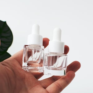 May include: Two small, clear glass bottles with square bases and white dropper caps. Each bottle features a glass pipette extending into the clear container, designed for liquid storage and precise dispensing in a compact, portable size.