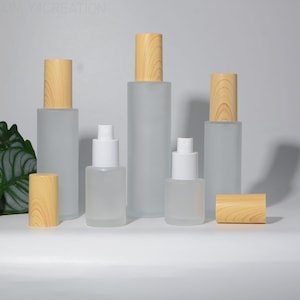 Puede incluir: Una colección de botellas de vidrio esmerilado con tapas de madera y dispensadores de bomba blancos. Las botellas varían en tamaño y forma, con un acabado de veta de madera natural en las tapas. Sugiere una línea de productos cosméticos.