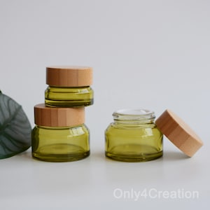 May include: Four small, olive-green glass jars with wooden lids. One jar is open, revealing a white substance inside. The jars are stacked and arranged on a white surface, suggesting a skincare or cosmetic product.