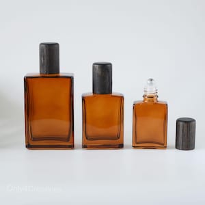 Könnte beinhalten: Drei bernsteinfarbene Parfümflaschen aus Glas mit dunklen Holzverschlüssen. Die Flaschen sind rechteckig mit abgerundeten Ecken. Eine Flasche hat einen Rollballaufsatz. Die Verschlüsse sind zylindrisch. Die Flaschen sind in einer Reihe vor weißem Hintergrund angeordnet.