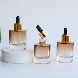 Op de afbeelding: Drie glazen druppelflessen met een bruine gradiënt. De flessen hebben goudkleurige kragen en zwarte druppelaars. Eén fles heeft de druppelaar verwijderd en de andere twee hebben de druppelaars ingebracht. De flessen staan op een wit oppervlak.