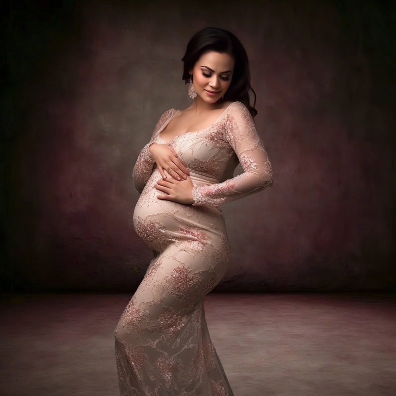 May include: A pregnant woman in a long, pink lace dress with a fitted bodice and long sleeves. She is standing in front of a dark brown background and is looking down at her stomach.