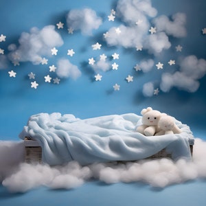 Op de afbeelding: Een witte rieten mand met een lichtblauwe deken erin. Twee witte knuffels, een beer en een konijn, zitten op de deken. De mand is omringd door witte wattenbolletjes, die lijken op wolken. De achtergrond is een lichtblauwe muur met witte sterren en wolken.