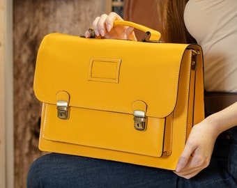May include: A yellow leather briefcase with brass clasps and a flap closure. The briefcase is held by a person wearing blue jeans.