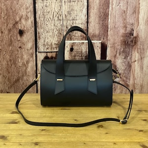 May include: A black handbag with a cylindrical body and a flap closure. The bag has two short handles and a long, adjustable shoulder strap. Gold-coloured metal accents are visible. The bag is on a wooden surface.