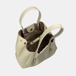 May include: Cream-colored leather handbag with two top handles and a detachable shoulder strap. The bag has a structured, rounded shape with a soft, textured exterior. Gold-toned hardware accents the bag, including the clasp and strap attachments.