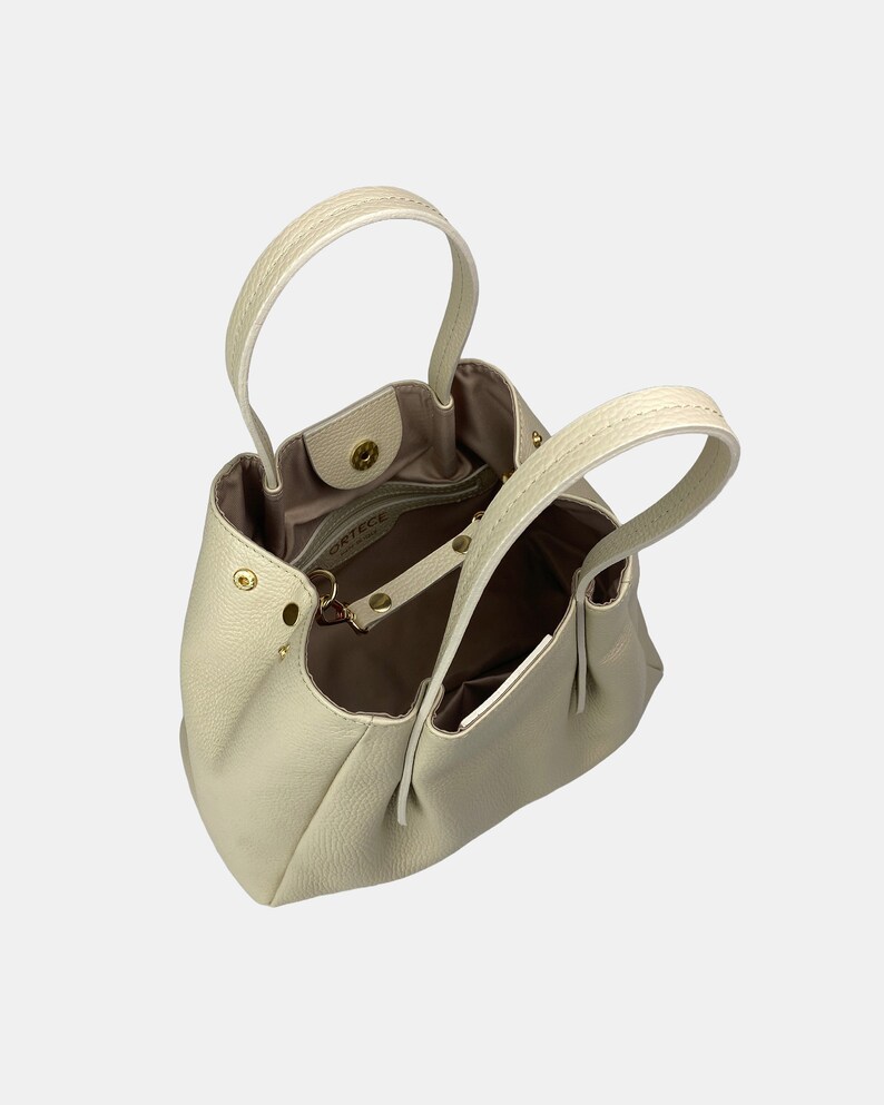 May include: Cream-colored leather handbag with two top handles and a detachable shoulder strap. The bag has a structured, rounded shape with a soft, textured exterior. Gold-toned hardware accents the bag, including the clasp and strap attachments.