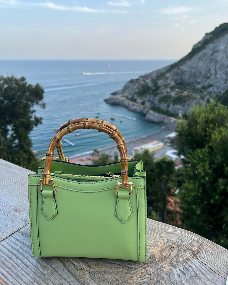 May include: A light green leather handbag with a bamboo handle. The bag is sitting on a wooden surface with a view of a coastline in the background.