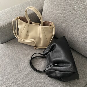 May include: Two leather handbags in cream and black rest on a gray sofa. The cream bag has a structured design with handles and a shoulder strap. The black bag is a smaller, gathered style with a single handle.