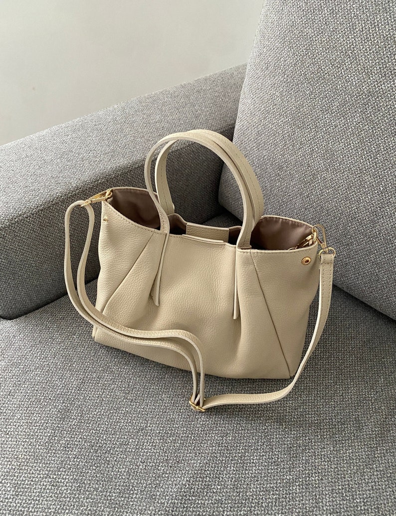May include: A cream-colored handbag with a textured exterior and a structured design. The bag features two short handles and a detachable shoulder strap. The interior lining is a contrasting brown color, and the hardware is gold-toned.