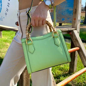 May include: A light green leather handbag with a bamboo handle. The bag has a top flap with a gold buckle closure.