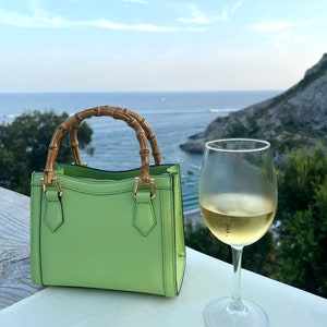 May include: A light green leather handbag with bamboo handles. The bag is sitting on a white table with a glass of white wine next to it. The background is a scenic view of the ocean and a rocky coastline.