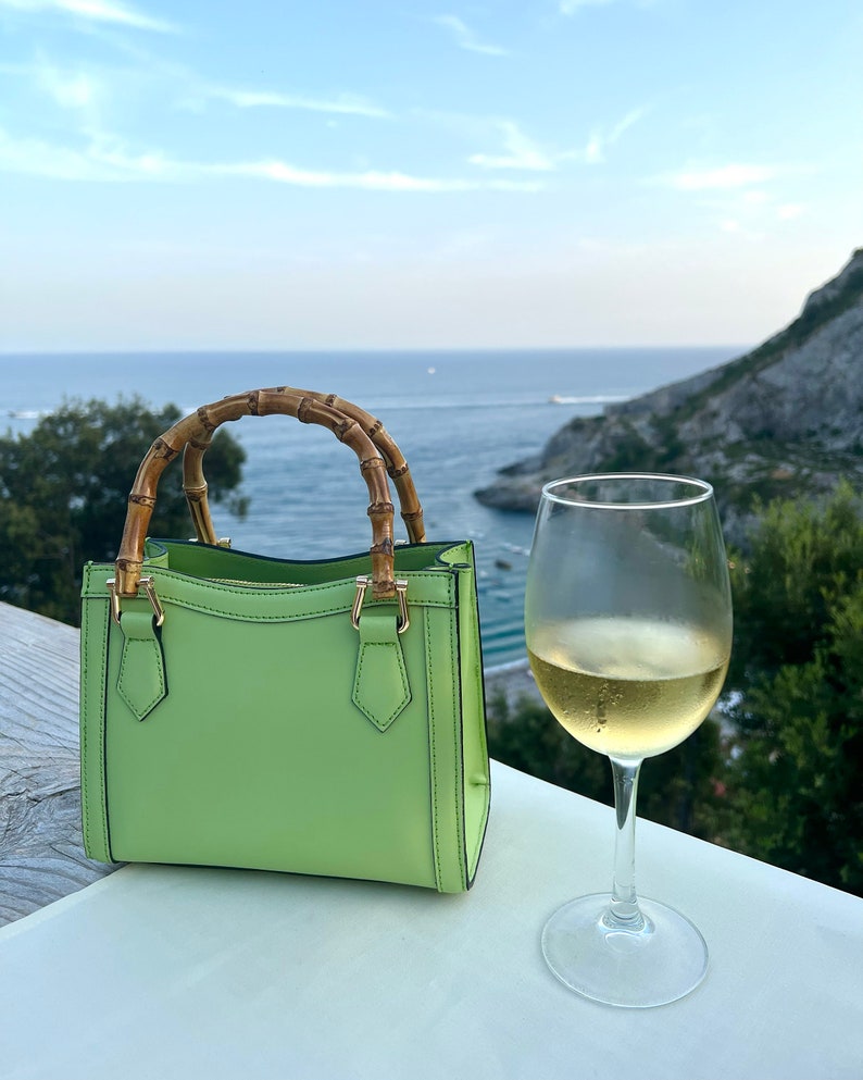 May include: A light green leather handbag with bamboo handles. The bag is sitting on a white table with a glass of white wine next to it. The background is a scenic view of the ocean and a rocky coastline.