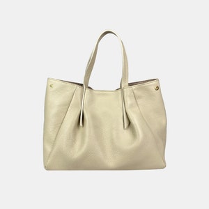 May include: A cream-colored leather tote bag with two handles. The bag has a gathered design on the front and sides, and gold-colored hardware accents. The interior lining is a darker shade of brown.