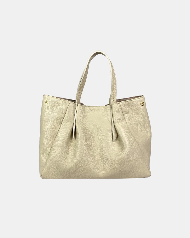 May include: A cream-colored leather tote bag with two handles. The bag has a gathered design on the front and sides, and gold-colored hardware accents. The interior lining is a darker shade of brown.
