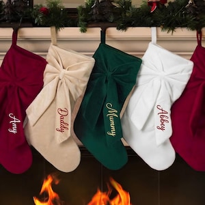 May include: Five velvet Christmas stockings hanging in a row, featuring a large decorative bow on the front of each. The stockings are coloured burgundy, beige, dark green, and white, with the names Amy, Daddy, Mummy, Abbey, and Aaron embroidered.