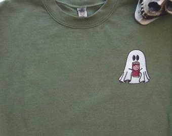 May include: An olive green crewneck sweatshirt featuring an embroidered design on the left chest. The embroidery depicts a white ghost holding a small red soda can. The sweatshirt has long sleeves with ribbed cuffs and a ribbed neckline.