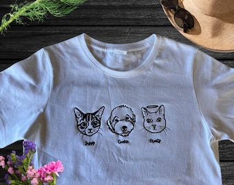 Puede incluir: Camiseta blanca con imágenes bordadas en negro de un gato, un perro y un gato con halo. Los nombres "Ziggy", "Cookie" y "Pruitt" están escritos debajo de las imágenes.