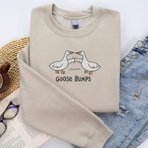 Może przedstawiać: Beżowa bluza z nadrukiem z kresk&oacute;wki przedstawiającym dwie białe gęsi zwr&oacute;cone do siebie, z napisem "Goose Bumps" poniżej. Gęsi są umieszczone nad falistą linią, sugerującą wodę. Bluza jest starannie złożona.