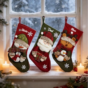 Puede incluir: Tres calcetines de Navidad colgados en una ventana. El izquierdo es rojo con un muñeco de nieve y el nombre Olivia. El central es verde con Santa Claus y el nombre Dad. El derecho es rojo con un reno y el nombre Mum. Incluyen copos de nieve.
