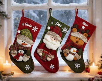 May include: Three Christmas stockings hanging in a window. The left is red with a snowman and the name Olivia. The center is green with Santa Claus and the name Dad. The right is red with a reindeer and the name Mum. Each features white snowflakes.