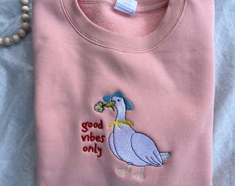 May include: A pink sweatshirt featuring an embroidered design of a goose wearing a blue hat and holding a flower in its beak. The text "good vibes only" is embroidered below the goose.