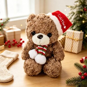 May include: A brown plush teddy bear with curly fur, wearing a red Santa hat with the name "Caitlin" embroidered in white, and a striped scarf in red, green, and white with two white pom-poms at the ends, featuring large black eyes.