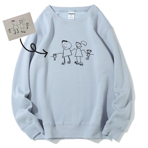 May include: A light blue crewneck sweatshirt featuring a black line drawing of a family on the front. The design depicts two adults and two children holding hands, matching the sketch shown on a small paper insert pointing to the graphic.