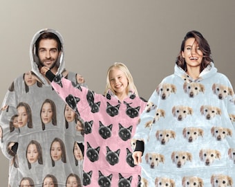 May include: Three hooded wearable blankets. One is light gray with repeating faces, another is pink with black cat faces, and the third is light blue with repeating dog faces. All have a soft, plush appearance and a large front pocket.