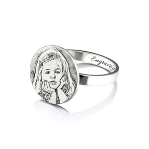 May include: A silver ring featuring a round, engraved photograph of a young girl. The ring is engraved with the word "Engrave" on the band.